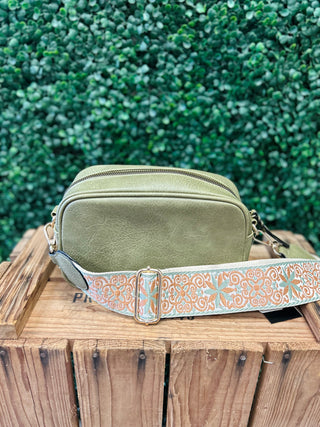 Olive green crossbody bag with decorative guitar strap sitting on wooden crate outdoors