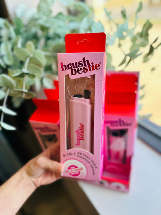 Hand holding pink Brush Bestie makeup brush in clear packaging with product boxes in background