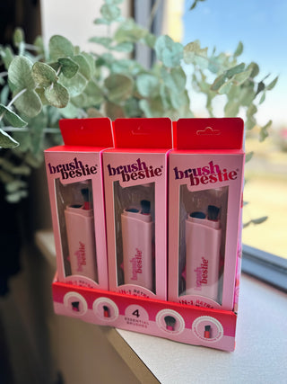 Three pink Brush Bestie makeup brush products in retail packaging displayed on a surface near eucalyptus plants.