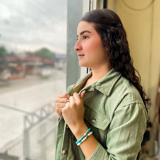 Woman wearing turquoise and white beaded Cabana Bracelet while looking out window in green corduroy shirt.
