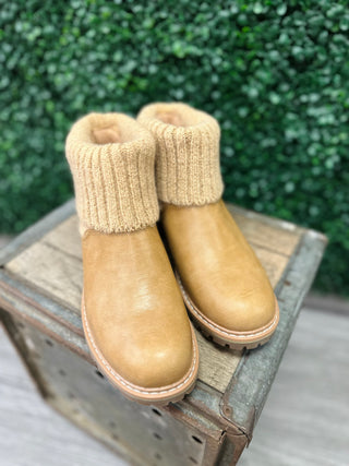 Caramel leather ankle boots with knit sweater cuffs displayed on wooden crate against green foliage background.