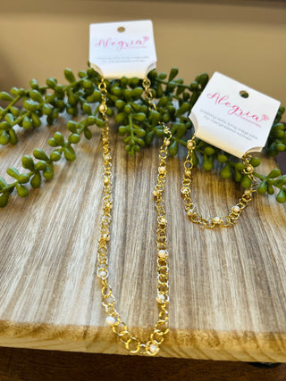 Gold chain bracelet by Alegria Accessories displayed on wooden surface with green foliage and product tags.