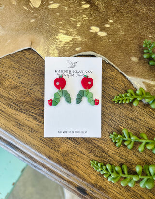 Handcrafted polymer clay caterpillar and apple dangle earrings displayed on a branded jewelry card.