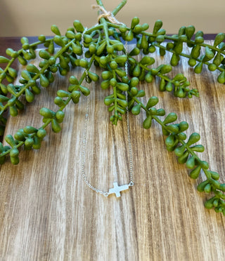 Silver cross pendant necklace on delicate chain displayed with green succulent plant on wood surface.