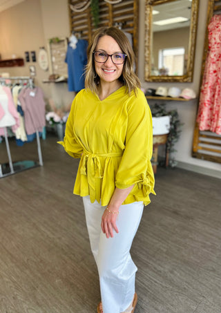 Woman wearing bright yellow hooded top with tie waist and white pants in clothing boutique store.
