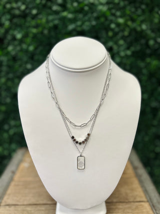 Layered silver chain necklace with beads and rectangular pendant displayed on white jewelry bust outdoors.