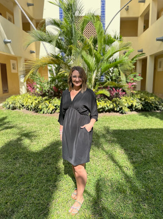 Woman wearing black wrap-style dress with three-quarter sleeves standing on grass in tropical resort setting.