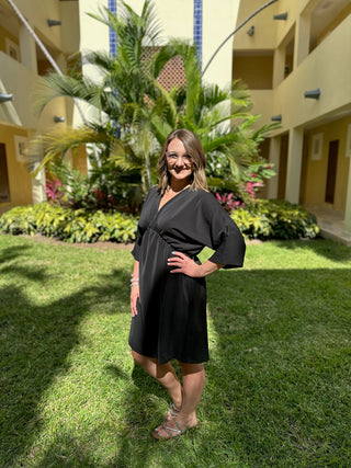 Woman wearing black wrap dress with three-quarter sleeves posing in tropical resort courtyard setting.