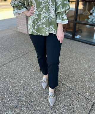 Woman wearing black stretch pants with green floral top and gray pointed-toe flats on sidewalk.