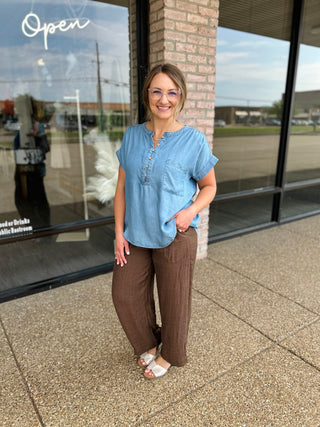 Woman wearing a relaxed short-sleeve chambray henley blouse with button placket and chest pocket.