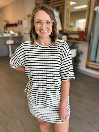Woman wearing black and white striped oversized top with glasses, smiling in boutique setting.