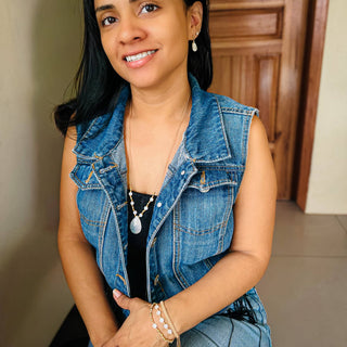 Woman wearing pearl drop necklace with denim vest, smiling indoors with wooden cabinet background.