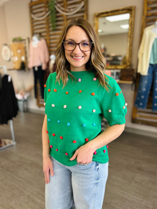 Woman wearing green knit sweater with colorful pom-pom details in clothing boutique setting