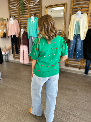 Woman wearing green knit sweater with colorful pom pom details and light blue jeans in boutique setting.