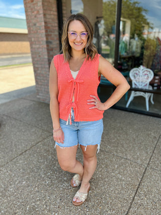Woman wearing coral sleeveless tie-front cardigan top with denim shorts, standing outdoors smiling.