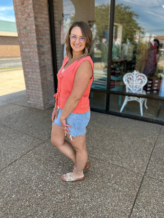 Woman wearing coral sleeveless cardigan top with denim shorts, standing outside storefront with brick columns.