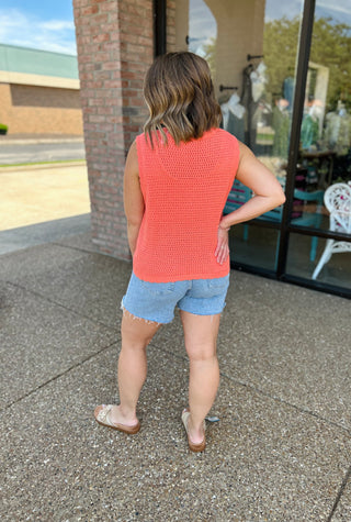 Woman wearing coral knit sleeveless cardigan top with denim shorts, viewed from behind in outdoor setting.