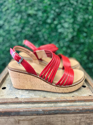 Red strappy wedge sandals with cork platform sole displayed on wooden surface with green foliage background.