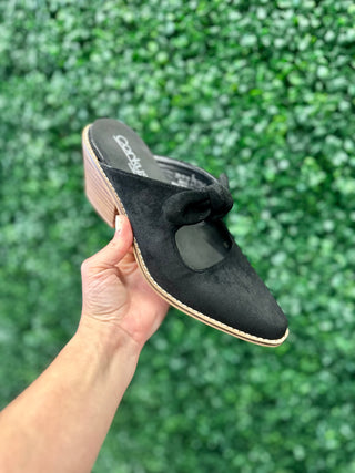 Corkys mule slide held up against green foliage backdrop, featuring suede upper with bow detail and wedge sole.