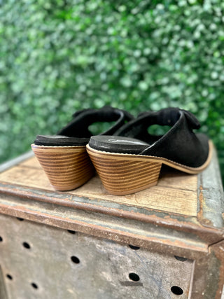 Corkys suede mule slides with stacked wood-look block heels and bow detail, displayed on rustic wooden crate.