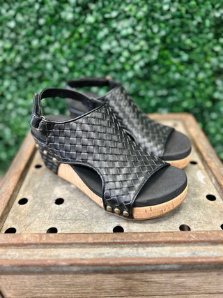 Black woven wedge sandals with cork platform and ankle strap displayed on wooden surface