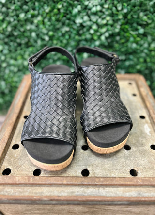 Black woven leather wedge sandals with cork sole and ankle strap on wooden surface