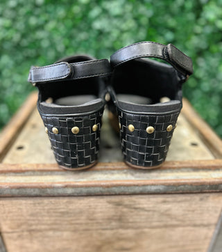 Black woven wedge sandals with ankle straps and gold studs displayed on wooden surface