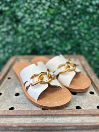 White leather sandals with gold chain detail on brown wooden surface with greenery background.