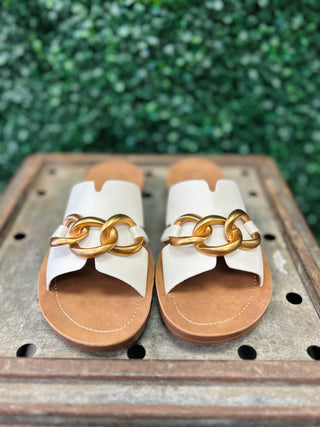 White slide sandals with gold chain detail on brown cork soles displayed on wooden surface.