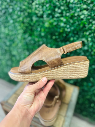Corkys Volta 2 leather wedge sandal with woven platform sole and slingback strap, held up against green foliage backdrop.