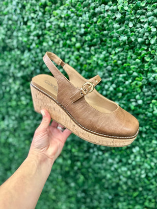 Corkys Pity Party leather buckle wedge sandal with cork platform, held against green foliage backdrop.