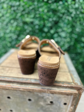 Corky's buckle wedge sandals with cork heel displayed on rustic wooden crate against greenery backdrop.