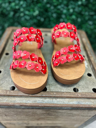 Red floral sandals with gold studs on wooden surface, featuring multiple flower-adorned straps and tan soles.