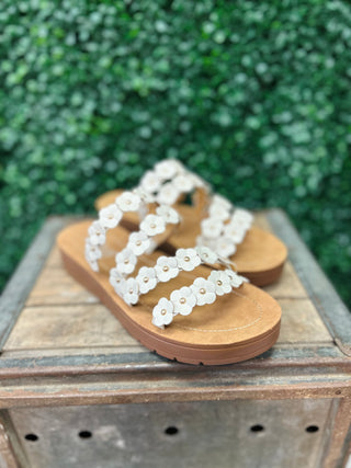 White floral sandals with flower embellishments and cork soles displayed on wooden surface outdoors.