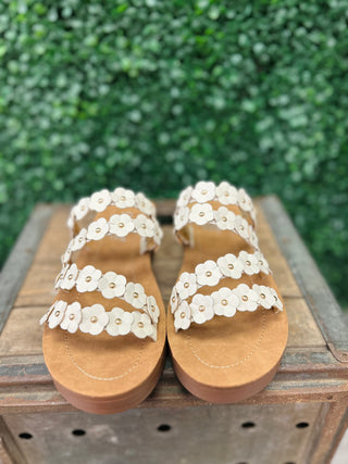 White floral sandals with decorative flower straps and cork soles displayed on wooden surface outdoors.