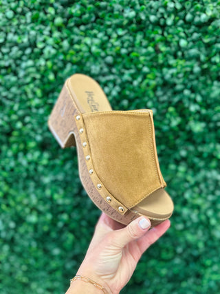 Corkys Gaslight suede wedge mule with cork platform and gold stud detailing, held against greenery backdrop.
