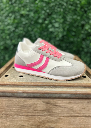 Grey and pink athletic sneakers with laces displayed on wooden surface with green foliage background.