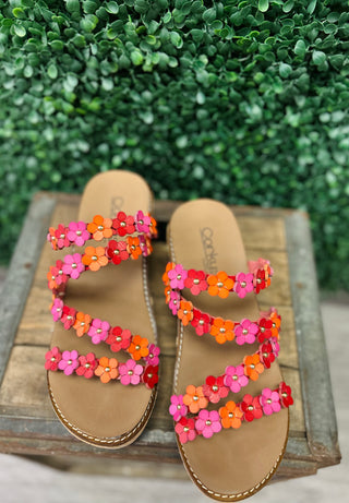 Colorful floral embellished wedge sandals with multiple straps displayed on wooden surface against green foliage background.