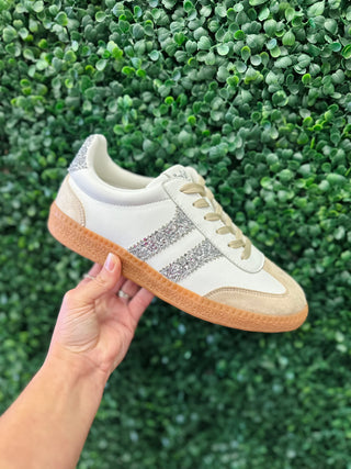 Corkys Heads Up Sneaker with glitter stripe detail and gum sole held against green foliage backdrop.