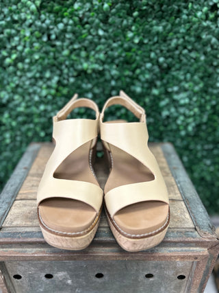Beige platform wedge sandals with ankle straps displayed on wooden surface with green foliage background