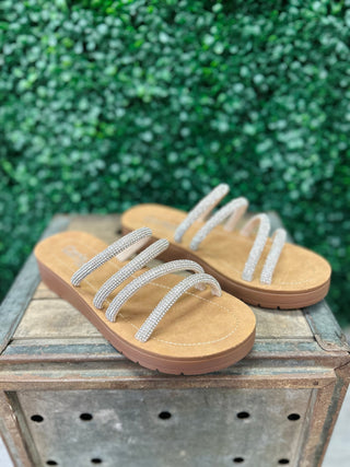 Corky's strappy rhinestone sandals with multiple embellished straps on wooden surface with green foliage background