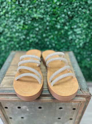 Corky's strappy rhinestone sandals with multiple embellished bands on wooden crate against green foliage background