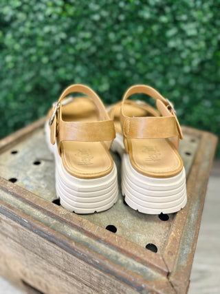 Caramel leather sandals with white chunky soles and adjustable straps displayed on weathered wooden crate outdoors.