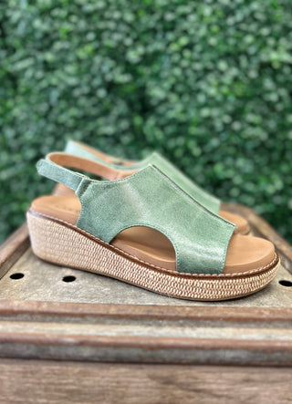 Green leather wedge sandal with woven sole and ankle strap on wooden surface