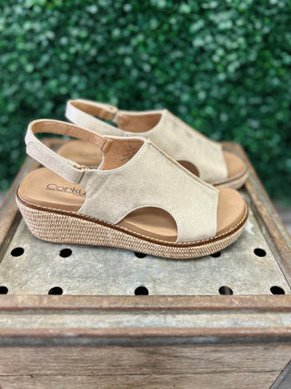 Gold metallic platform wedge sandals with ankle strap displayed on wooden surface with greenery background.