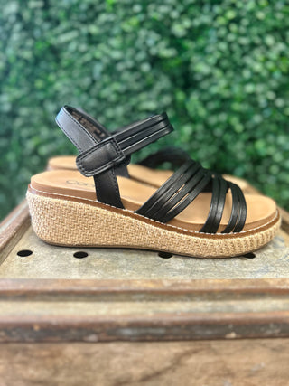 Black strappy wedge sandal with woven platform sole displayed on wooden surface with greenery background.