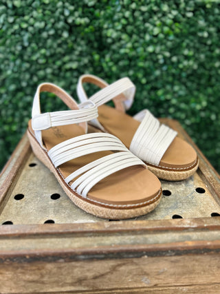 White strappy wedge sandals with cork soles displayed on wooden surface with green foliage background.