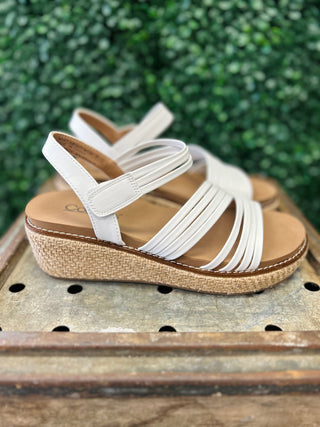 White strappy wedge sandals with cork platform sole displayed on wooden surface with green foliage background.