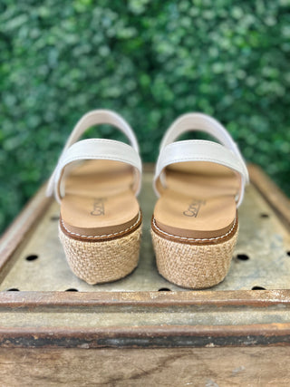 White wedge sandals with espadrille platform soles and ankle straps displayed on wooden surface outdoors.