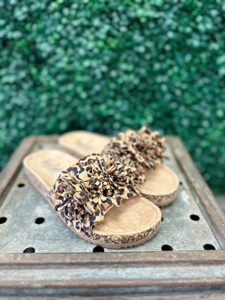 Corky's Wild Child leopard print sandals with ruffled straps on wooden surface with green foliage background